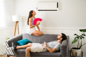 Woman looking at broken AC in despair while fanning herself and man lying on couch exhausted from the heat.