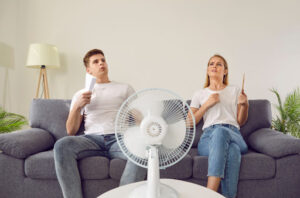 A man and woman sitting on a couch with a portable fan while they fan themselves with pieces of paper.